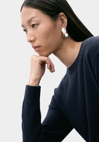 Silverfärgade abstrakta örhängen med en blank yta. Modellen bär en mörkblå stickad tröja. Mjuk textur och profilbild i närbild.