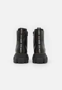 Bottes basses en cuir noir avec semelles épaisses et languettes en tissu de marque portant l'inscription "Steve Madden" à l'arrière.