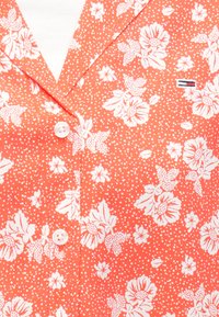 Czerwona koszula z krótkim rękawem w kwiaty z białymi wzorami kwiatów i małymi białymi kropkami. Posiada zaostrzony kołnierzyk i zapięcie na guziki na środku.