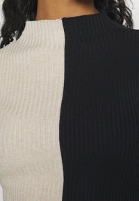 Närbild av en räfflad, tvåfärgad tröja med beige på vänster sida och svart på höger sida, bärs av en person med mörkt, lockigt hår.