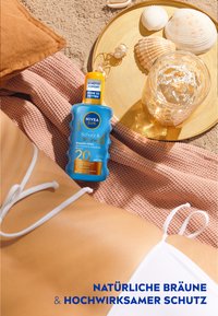 NIVEA Sun sunscreen bottle with a blue body and yellow cap on tan fabric; nearby glass, seashells, and textured surfaces create a beach scene.