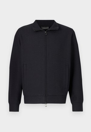 Veste noire zippée avec un motif texturé, col montant, poches latérales et poignets côtelés. Confectionnée dans un tissu doux et tissé.