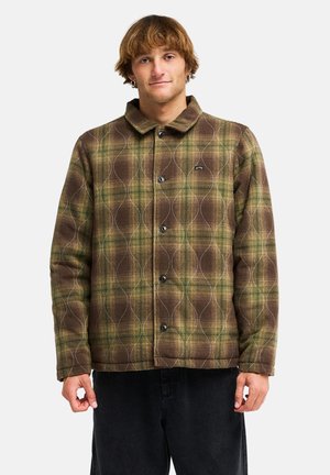 Junger Mann trägt grün-braun karierte gesteppte Jacke mit schwarzen Knöpfen und schwarze Hose, steht vor weißem Hintergrund.