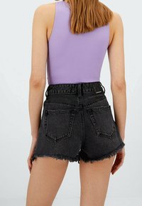 Zwarte denim shorts met rafelige zoom, hoge taille en een stoffen riem. Het model draagt een lila mouwloze top die het ontwerp op de rug laat zien.