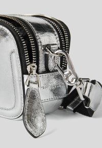 Angolo di una borsa in pelle argentata con texture, cerniere nere, una chiusura argentata e una tracolla attaccata su uno sfondo grigio chiaro.