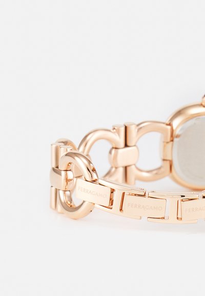 Rose gold metal watch with linked chain bracelet and circular face. Features engraved brand name on several links. Smooth, polished surface.