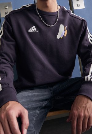 Personne portant un sweat-shirt Adidas foncé avec des bandes blanches sur les manches, un collier chaîne argenté et un jean foncé, assise devant des casiers.