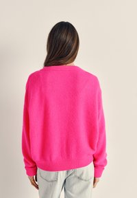 Sieviete ar gariem brūniem matiem valkā spilgti rozā džemperi un gaiši zilas džinsas, stāvot pret vienkāršu bēša fona sienu.