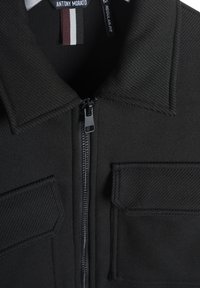 Zwarte jas met een stevige textuur, voorzien van een ritsluiting en twee voorzakken. Label geeft aan "ANTONY MORATO REGULAR-FIT."