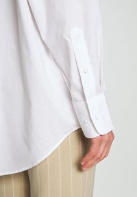 Chemise blanche à manches longues avec un col pointu, des poignets boutonnés et un ourlet incurvé ; tissu en coton lisse avec un éclat subtil.