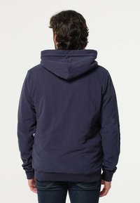 Marineblauwe hoodie gemaakt van zachte stof, met een grote capuchon, geribbelde manchetten en een ontspannen pasvorm. De achterkant toont een gladde textuur en een eenvoudig ontwerp.
