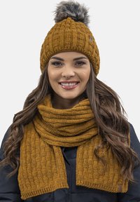 Conjunto de gorro y bufanda en color mostaza. El gorro presenta un patrón texturizado con un pompón de piel sintética. La bufanda tiene un diseño de punto similar.