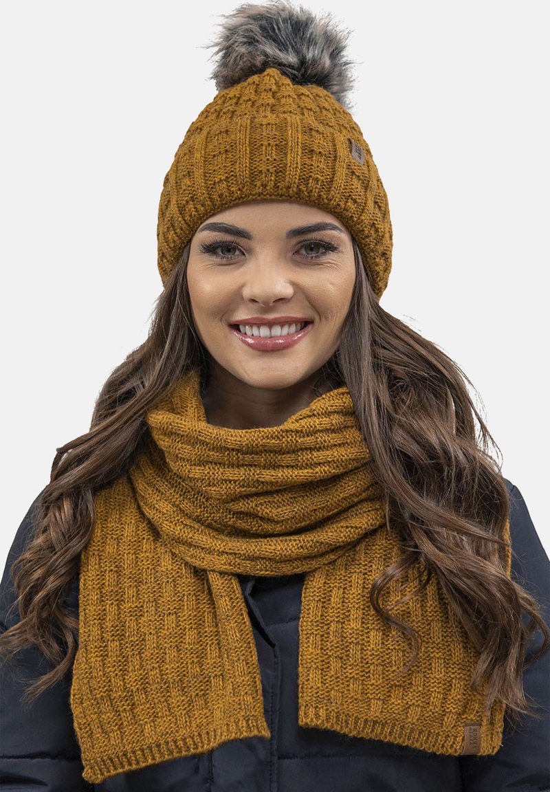 Conjunto de gorro y bufanda en color mostaza. El gorro presenta un patrón texturizado con un pompón de piel sintética. La bufanda tiene un diseño de punto similar.