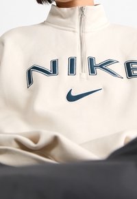 Beige sweatvest met rits en een hoge kraag, voorzien van blauwe Nike-merknaam en logo aan de voorkant, gemaakt van zacht, gestructureerd textiel.