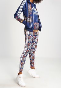 Marinblå sportoverall med blommönster; zip-up-hoodie och figursydda byxor med tre vita ränder på sidorna. Vita sneakers kompletterar outfiten.