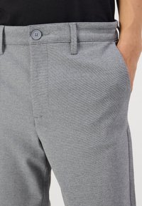 Graue Hosen aus flach gewebtem Stoff, mit Knopfverschluss, seitlichen Taschen und tailliertem Schnitt mit Gürtelschlaufen.