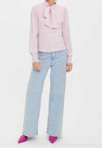 Blusa a maniche lunghe rosa chiaro con colletto a fiocco e tessuto testurizzato, abbinata a jeans a gamba larga blu chiaro e scarpe con punta a destra rosa brillante.