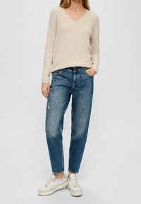 Donna che indossa un maglione beige a V, jeans blu comodi e scarpe da ginnastica bianche, in piedi con una mano in tasca su uno sfondo neutro.