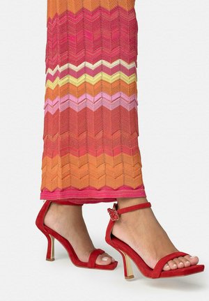 Sandalias de tacón alto de ante rojo con tiras en el tobillo, usadas con una falda de punto con zigzag multicolor en tonos naranja, rosa, amarillo y rojo.
