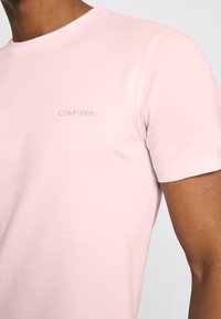 Camiseta de algodón de color rosa claro con cuello redondo. Presenta un sutil logotipo "Calvin Klein" en tonos similares en el lado izquierdo del pecho. Textura suave, forma ajustada.