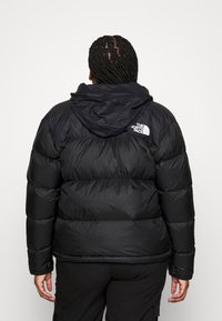 Čierna pérová bunda s kapucňou, má prešívané časti a elastické manžety. Logo značky je umiestnené na hornej časti chrbta.