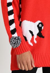 Maglione rosso lavorato a maglia con un design di cane bianco e nero sul davanti e una trama a righe bianco e nero sulle maniche, completato da un pompon.