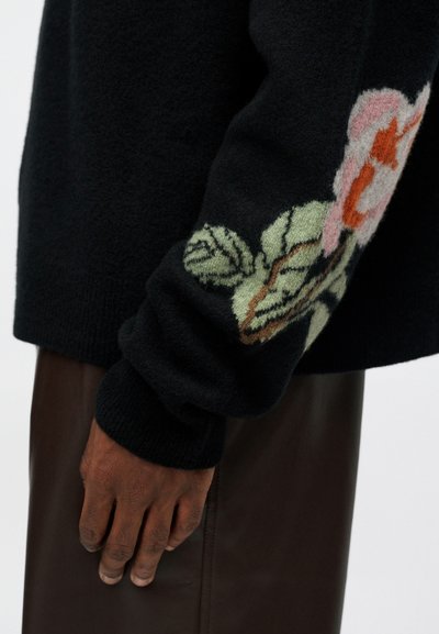 O mână cu pielea închisă la culoare, odihnindu-se pe lângă corp, îmbrăcată într-un pulover negru cu broderie florală roz și verde pe mânecă și în pantaloni de piele maro.