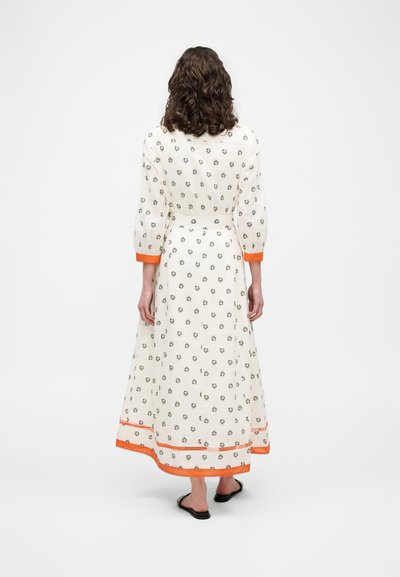Femme aux cheveux bouclés portant une longue robe blanche à petits imprimés floraux, avec des poignets et un ourlet orange, assortie à des sandales noires sans lacets, vue de dos.