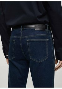 Tamno plavi traperice imaju uski kroj, dva stražnja džepa s kutnim preklopima i kožni crni detalj na pojasu.