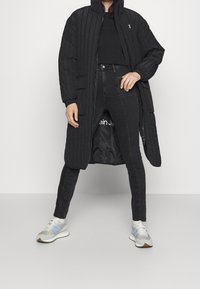 Abrigo largo negro acolchado con cuello alto, usado sobre una blusa negra y jeans negros. Presenta bolsillos laterales y una cremallera expuesta. Zapatillas grises con detalles azules.