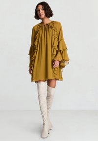 Olivgrünes Kleid mit gerafften Ärmeln und Ausschnitt, verfügt über eine Schnürung, kombiniert mit weißen kniehohen Stiefeln. Glatter, leichter Stoff.