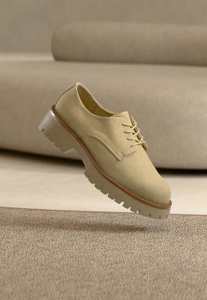 Beige suède veterschoen met dikke zool zweeft boven een getextureerd tapijt, neutraal gekleurde bank op de achtergrond. Geen mensen aanwezig.