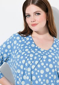 Lichtblauwe blouse met witte stippen, V-hals en geplooid detail aan de voorkant. Zachte stof met een gladde textuur en driekwartmouwen.