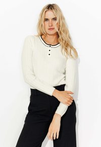 T-shirt long à manches blanches côtelé avec un col noir et une patte de boutonnage à trois boutons, associé à un pantalon noir ajusté. Détail de tissu texturé visible.