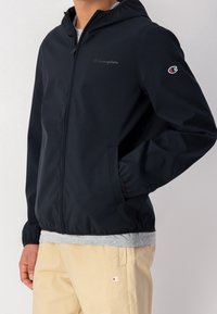 Marinblå kapuschongjacka i mjukt material med framdragkedja, elastiska muddar och Champion-logotypen på bröstet och ärmen.