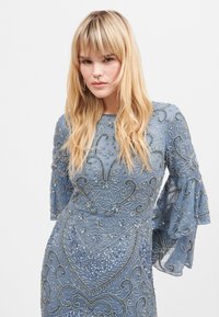 Niebieska sukienka z koralikami i długimi, rozszerzanymi rękawami. Projekt zawiera wzory kwiatowe i cekiny, co nadaje jej teksturowany wygląd.