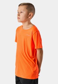 Jongen met kort haar, gekleed in een oranje kortemouwshirt en donkere broek, zijwaarts staand tegen een effen achtergrond.