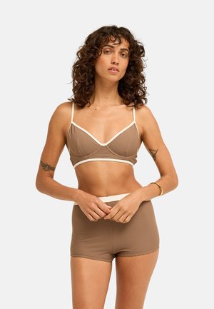 Donna con capelli ricci che indossa un bralette marrone a coste e pantaloncini coordinati a vita alta, in piedi con le mani giunte davanti.
