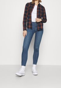 Camicia in flanella a scacchi blu e arancione, maglia bianca aderente, jeans skinny blu e sneakers bianche con suola texture.
