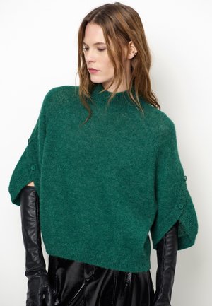 Jonge vrouw draagt een losse groene gebreide trui met knoopsluitingen aan de mouwen en lange zwarte leren handschoenen, kijkt naar beneden en opzij.