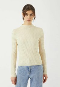 Kvinna som bär en krämfärgad texturerad långärmad topp och ljusblå jeans, stående mot en enkel vit bakgrund.