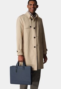 Beiger Trenchcoat mit schwarzen Knöpfen und einer navyblauen Lederaktentasche. Die Aktentasche verfügt über Griffe und eine glatte Textur. Maßgeschneiderte graue Hose.