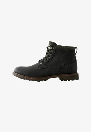 Black leather ankle boot with green textured collar, metal eyelets, and brown striped sole. Features a pull tab on the back for easy wear.