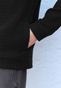 Melnā teksturētā džemperī ar brīvu piegriezumu, tajā ir sānu kabata un aproces uz piedurknēm. Materiāls izskatās mīksts un austs.