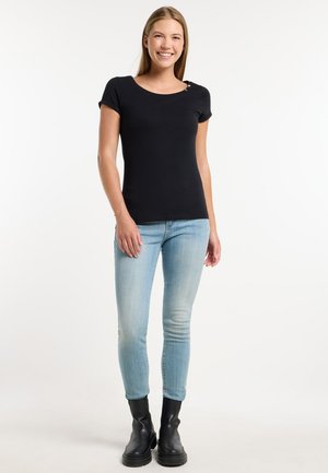 Joven mujer sonriente de pie con las manos relajadas, vistiendo camiseta negra de manga corta, vaqueros azul claro y botas negras hasta el tobillo sobre fondo liso.