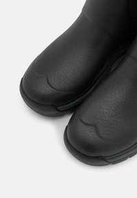 Mustat vettä hylkivät saappaat tekstuuripinnalla, pyöristetyillä varpailla ja tukevalla pohjalla. Suunnittelu on tyylikäs ja käytännöllinen. Ei näkyvää metalliosaa.