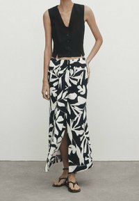 Zwart mouwloos topje met knoopdetail, gecombineerd met een lange, zwart-witte rok met bloemmotief, voorzien van een zijsplit en platte sandalen.