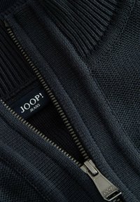 Zbliżenie ciemnego dzianinowego ubrania z częściowo otwartym metalowym zamkiem błyskawicznym, odsłaniającym czarną metkę z białym napisem "JOOP! JEANS".