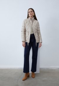 Donna che indossa una giacca trapuntata beige, top verde chiaro, jeans blu scuro e stivaletti marroni con tacco, in piedi contro un muro bianco.