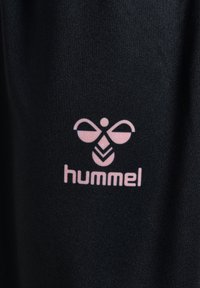 Czarna tkanina o gładkiej fakturze zawiera różowe logo Hummel, składające się z stylizowanej ikony pszczoły, znajdującej się nad nazwą marki.